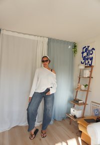 Witte off-shoulder gebreide trui, blauwe jeans en zwarte teenslippers. Natuurlijke houten vloer, lichte achtergrond met gordijnen en een decoratieve plank.