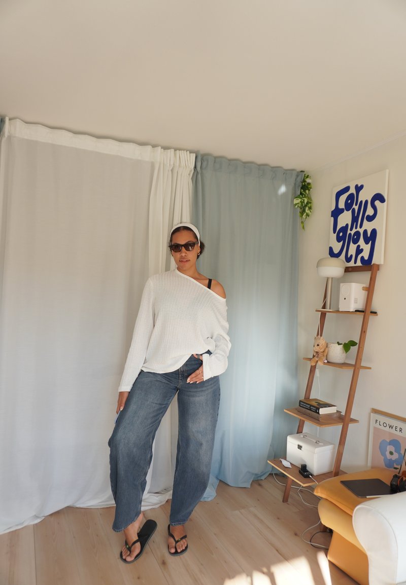 Bianco maglione a spalla scoperta, jeans blu e infradito nere. Pavimento in legno naturale, sfondo chiaro con tende e mensola decorativa.