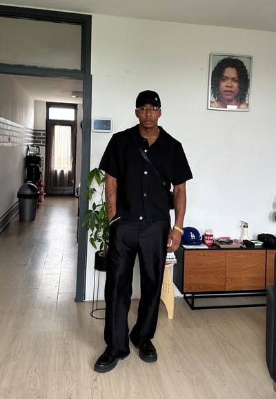 Camisa negra de punto de manga corta con botones, combinada con pantalones anchos negros y zapatillas negras. La habitación presenta un suelo de madera y plantas.