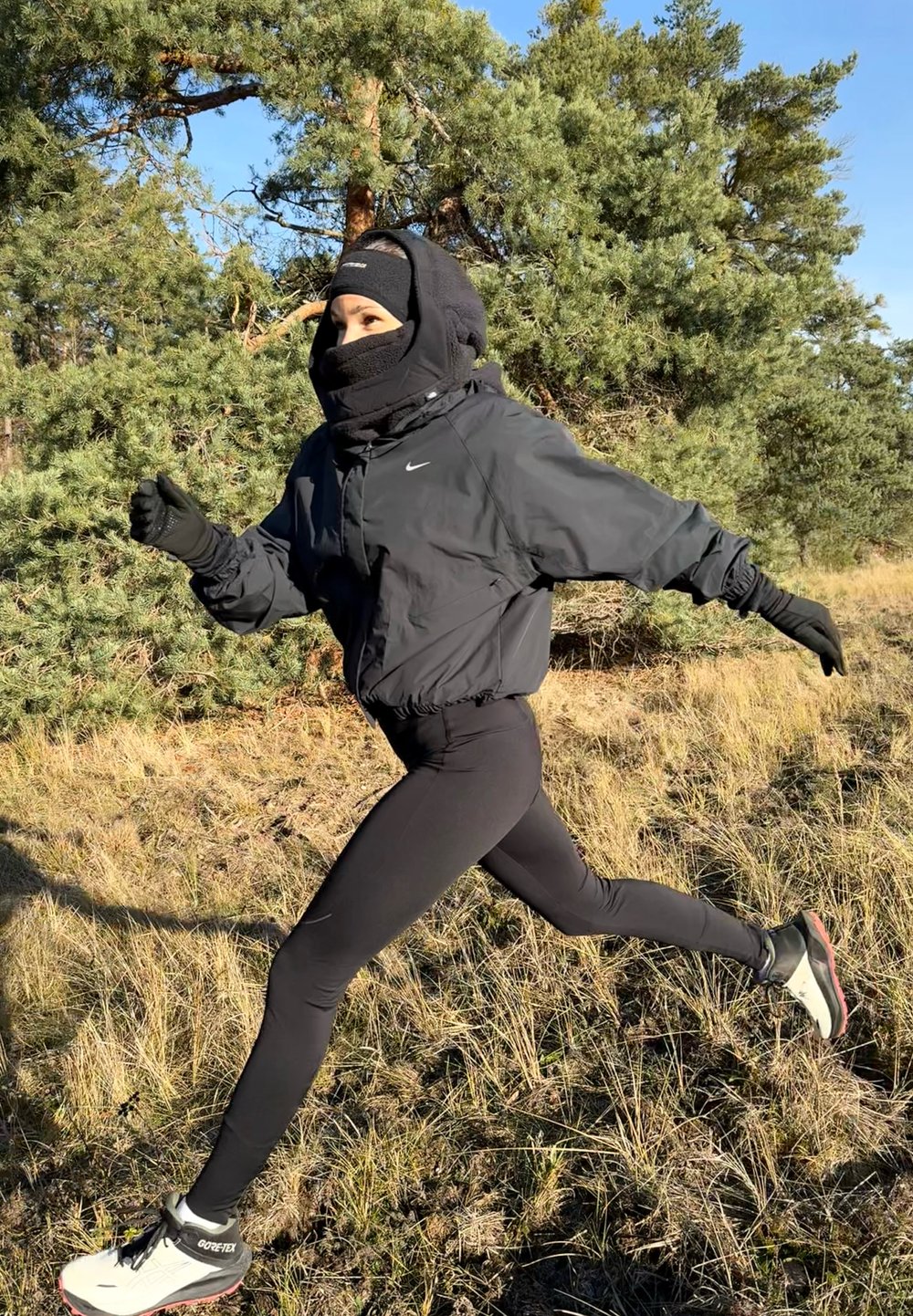 Giacca atletica nera, leggings neri e scarpe da corsa invernali; coperta con una maschera per il viso e guanti, corre su un terreno erboso.