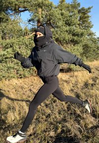 Giacca atletica nera, leggings neri e scarpe da corsa invernali; coperta con una maschera per il viso e guanti, corre su un terreno erboso.