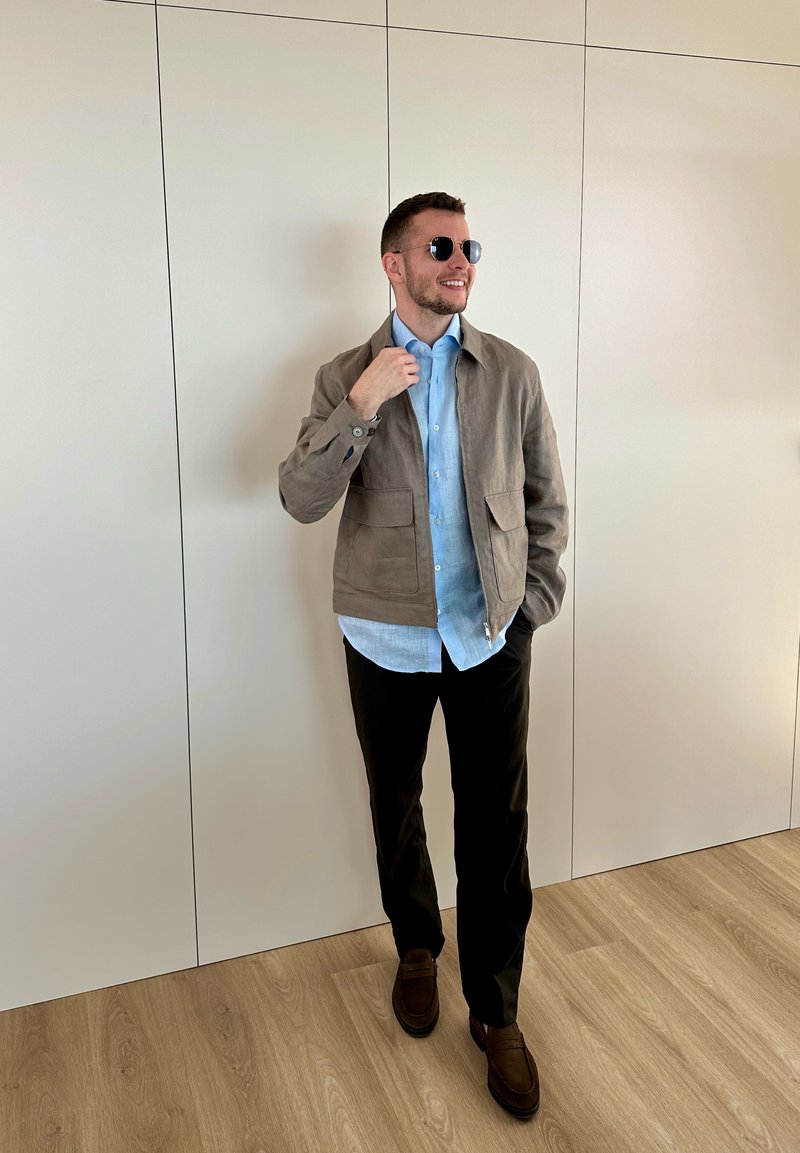 Hombre con gafas de sol, camisa azul clara, chaqueta beige, pantalones negros y zapatos marrones, de pie en el interior frente a una pared blanca con paneles.