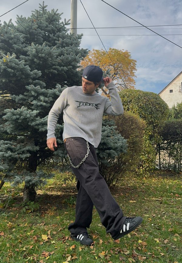 Pulover gri cu mâneci lungi și design grafic, pantaloni negri largi și adidași negri cu dungi albe. Conține un accesoriu tip lanț. Fond cu verdeață.