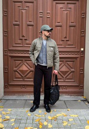 Chaqueta acolchada oliva, suéter gris, pantalones negros y zapatos negros. Sosteniendo una bolsa negra. Puerta decorativa marrón y hojas amarillas esparcidas en primer plano.