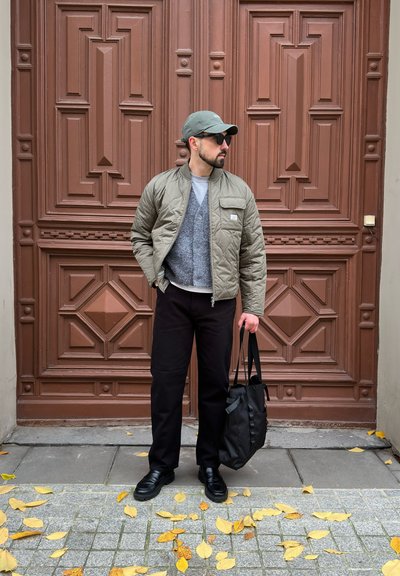 Chaqueta acolchada oliva, suéter gris, pantalones negros y zapatos negros. Sosteniendo una bolsa negra. Puerta decorativa marrón y hojas amarillas esparcidas en primer plano.