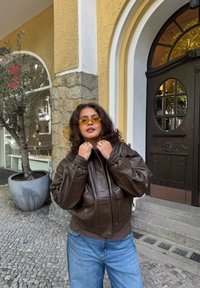Geacă bomber maro din piele, cu design scurt și manșete elastice, combinată cu blugi din denim bleu deschis. Ochelari de soare galbeni completează ținuta.