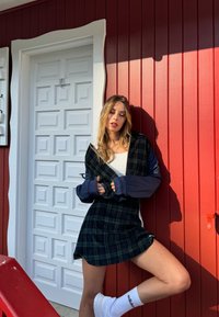 Πλαϊνό φόρεμα σε σκούρο πράσινο και μαύρο, επικαλυμμένο με μπλε τζάκετ. Λευκά sneakers και κάλτσες συμπληρώνουν το σύνολο, με φόντο ένα κόκκινο ξύλινο τοίχωμα.