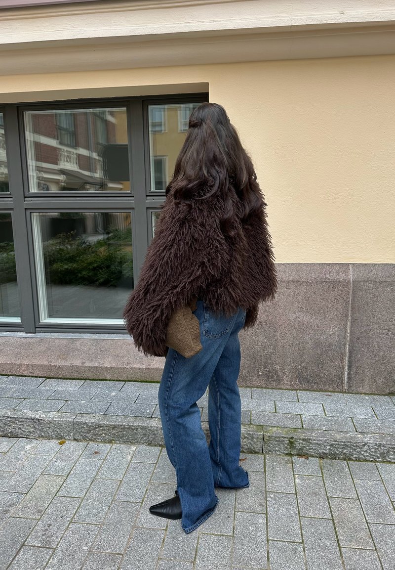 Kosmata rjava prevelika jakna, modre razširjene hlače in črni koničasti škornji. Ženska stoji ob stavbi z velikimi okni.