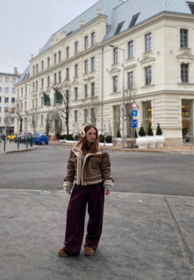Chaqueta de borreguito marrón con forro crema, combinada con pantalones sueltos de color púrpura oscuro. El escenario presenta un edificio histórico en el fondo.
