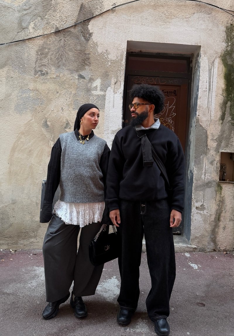 Deux personnes se tiennent devant un mur texturé ; l'une porte un gilet gris et un foulard noir, l'autre porte des lunettes et une tenue noire.