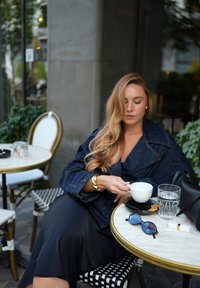 Džinsinė striukė, juoda sijonas, laiko baltą kavos puodelį, su juoda rankine ir apvaliais saulės akiniais ant marmurinio stalo. Fone žalios lapijos.