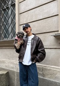Veste bomber en cuir marron avec poignets côtelés, t-shirt blanc et jean bleu ; tenant un petit chien vêtu d'un manteau rose, debout près d'un mur texturé.