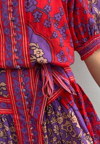 Rochie florală roșie și mov, cu mâneci bufante, butoane decorative și o curea legată. Prezintă modele îndrăznețe și țesătură texturată.