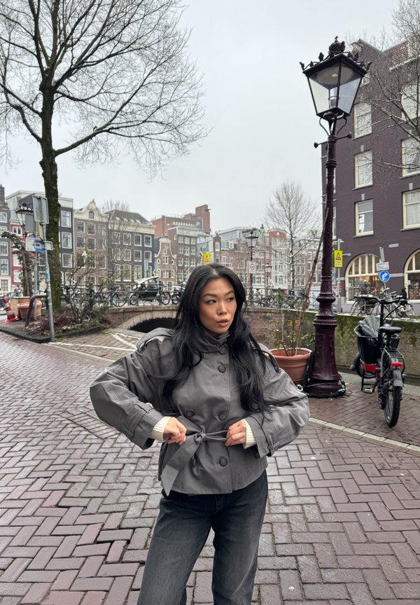 Femme ajustant sa ceinture sur une veste grise, debout sur une rue pavée près d'un canal avec des vélos et des bâtiments traditionnels hollandais par temps couvert.