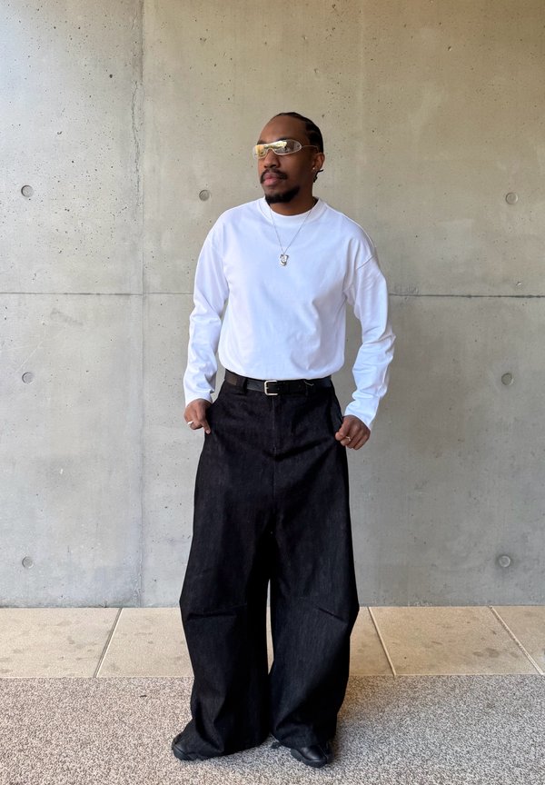 Homme aux cheveux tressés portant une chemise blanche à manches longues, un pantalon noir à jambes larges, des lunettes de soleil et un collier argenté, debout contre un mur en béton.