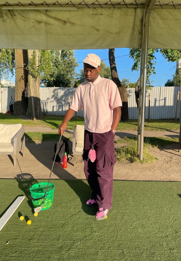 Camisa polo rosa, calças cargo roxas escuras com um pompom rosa, calçado rosa. Cesta verde segura bolas de golfe amarelas sobre uma superfície de relva.