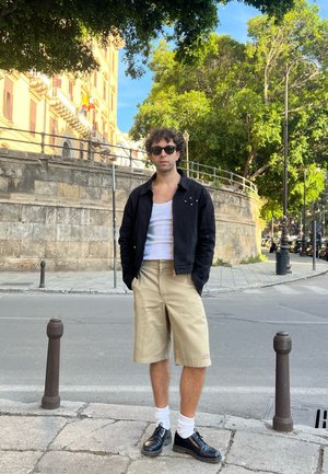 Chaqueta negra con detalles plateados sobre una camiseta sin mangas blanca, pantalones cortos beiges, calcetines blancos y zapatos negros, con un fondo urbano.