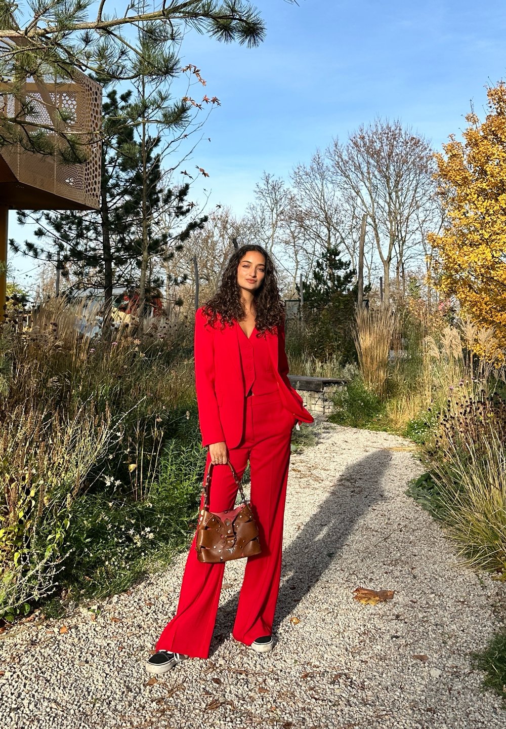 Sarkans pielāgots uzvalks ar pieguļošu žaketi un plašām biksēm. Apvienots ar brūnu somiņu ar metāliskām detaļām. Āra dārza vidē.