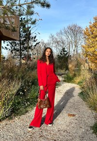 Sarkans pielāgots uzvalks ar pieguļošu žaketi un plašām biksēm. Apvienots ar brūnu somiņu ar metāliskām detaļām. Āra dārza vidē.