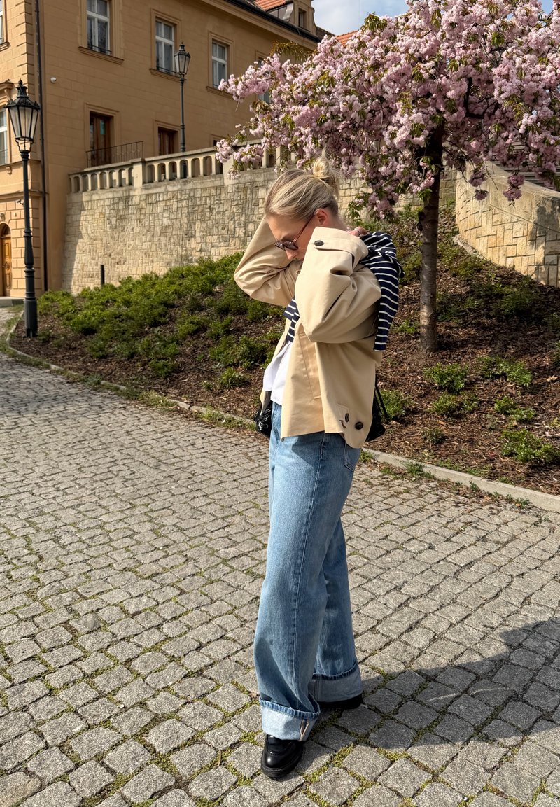 Femme ajustant sa veste debout dans une rue pavée près d'un arbre en fleurs et d'un bâtiment en pierre historique en pleine journée.