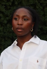 Biała koszula z kołnierzykiem i małym wyszywanym logo na piersi, zestawiona ze złotymi kolczykami kołami i naturalnymi włosami na tle zielonych liści.