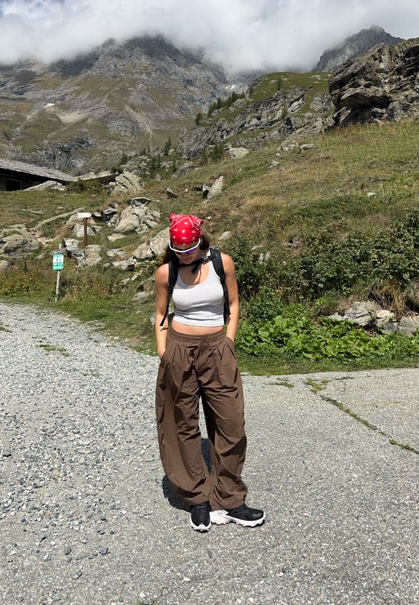 Giovane donna con bandana rossa, canotta grigia, pantaloni marroni e scarpe da ginnastica nere in piedi su un sentiero di ghiaia con montagne rocciose e vegetazione sullo sfondo.
