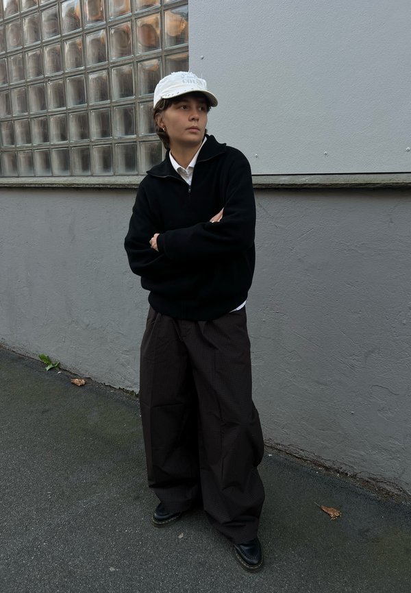 Persona joven vestida con una gorra blanca, suéter negro con cremallera, pantalones anchos oscuros y zapatos negros, de pie con los brazos cruzados frente a una pared gris.