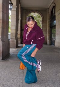 Bordeaux fleece pullover med zipkrave, teal træningsbukser med pink striber og sorte sneakers med multicolore accenter. Orange taske synlig.