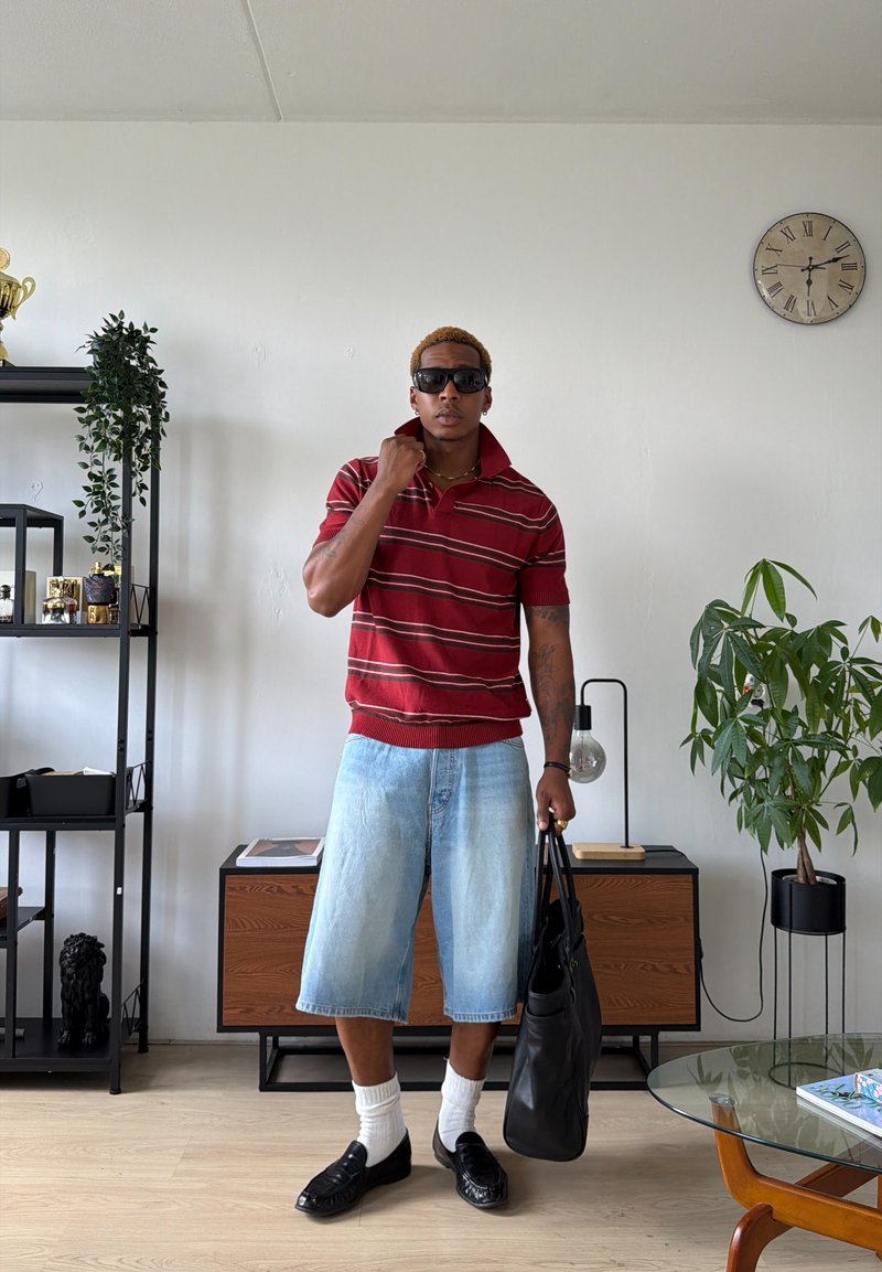 Homme portant des lunettes de soleil, polo rayé rouge, short en jean clair, chaussettes blanches, chaussures noires, tenant un sac noir dans une pièce moderne avec des plantes et une horloge.