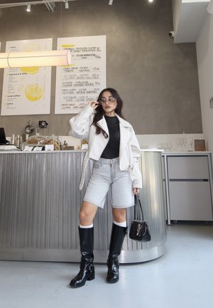 Mujer ajustándose las gafas de sol, de pie en un café moderno, vestida con chaqueta blanca, top negro, pantalones cortos vaqueros, botas negras y sosteniendo un bolso negro.