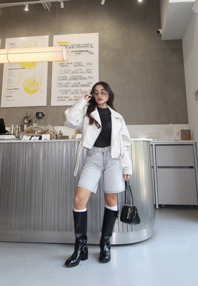 Mujer ajustándose las gafas de sol, de pie en un café moderno, vestida con chaqueta blanca, top negro, pantalones cortos vaqueros, botas negras y sosteniendo un bolso negro.