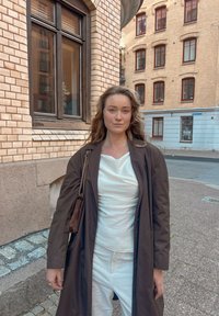 Abrigo largo marrón sobre una blusa blanca texturizada y pantalones blancos. Un pequeño bolso oscuro con correa de cadena. Muro de ladrillo y ventanas al fondo.