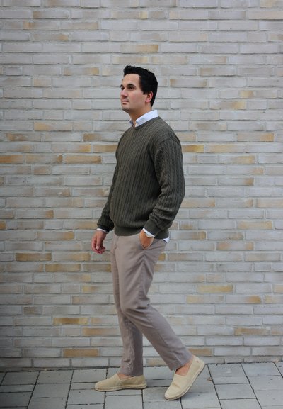 Suéter de punto en verde oscuro sobre una camisa blanca de cuello, combinado con pantalones beiges claros y espadrilles en tono beige, de pie frente a una pared de ladrillo.