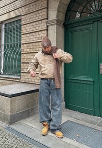 Bežna kapuca jakna preko rjave marame, plavi široki traperice i bež čizme, stoji ispred zeljenih vrata sa pozadinom od ciglastog zida.