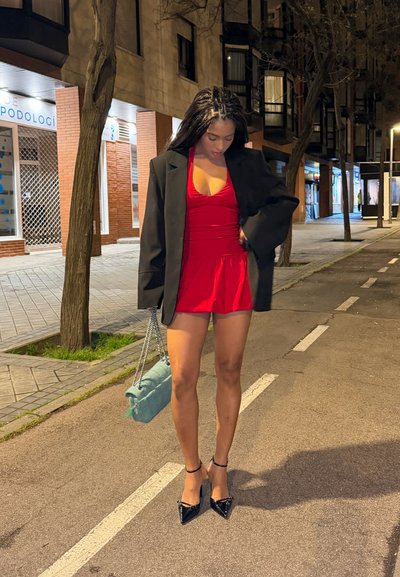 Mujer con vestido rojo y blazer negro sosteniendo un bolso azul claro, de pie en una calle de la ciudad por la noche con árboles y edificios cerca.