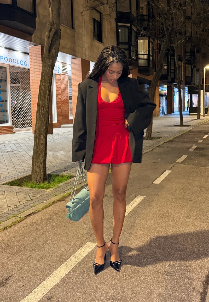 Woman in red dress and black blazer holding a light blue handbag, standing on a city street at night with trees and buildings nearby.