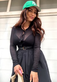 Top nero a coste con bottoni abbinato a una gonna pieghettata nera. Indossa un berretto verde e tiene una borsa metallica. Capelli lunghi castani.