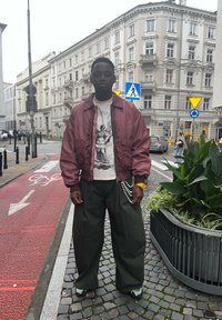 Bordowa kurtka bomber, graficzna biała koszulka, oversize'owe zielone spodnie, żółty zegarek, perłowy łańcuszek oraz czarne buty na stonowanym tle miejskim.