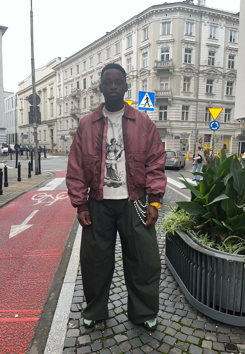 Bordeauxfarbene Bomberjacke, grafisches weißes T-Shirt, übergroße grüne Hose, gelbe Uhr, Perlenkette und schwarze Schuhe vor einem gedämpften Stadt-Hintergrund.