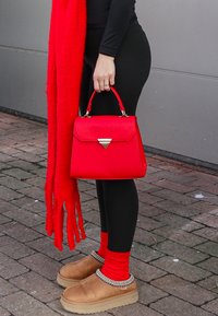 Sac à main rouge avec un accent métallique triangulaire, surface texturée. Assorti à des leggings noirs, une écharpe rouge et des chaussures beiges avec des bordures décoratives.