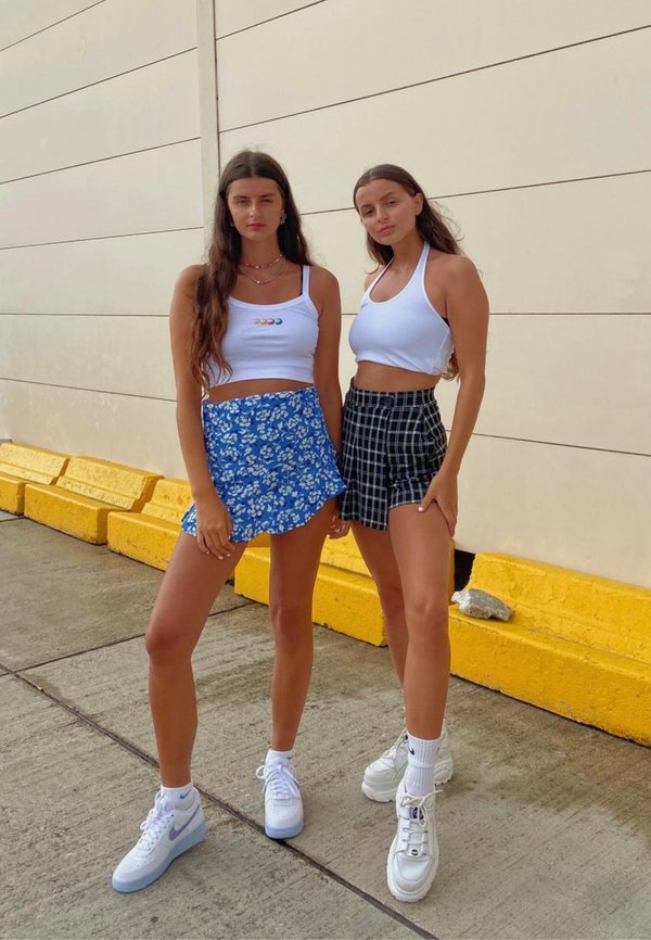 Dos personas están de pie una al lado de la otra. Una lleva un top corto blanco y una falda floral azul; la otra lleva un top corto blanco y una falda a cuadros negros. Ambas llevan zapatillas.