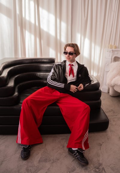 Joven con gafas de sol, chaqueta negra, pantalones anchos rojos y bufanda roja alrededor del cuello sentado en un sofá moderno negro en una habitación luminosa con cortinas blancas.