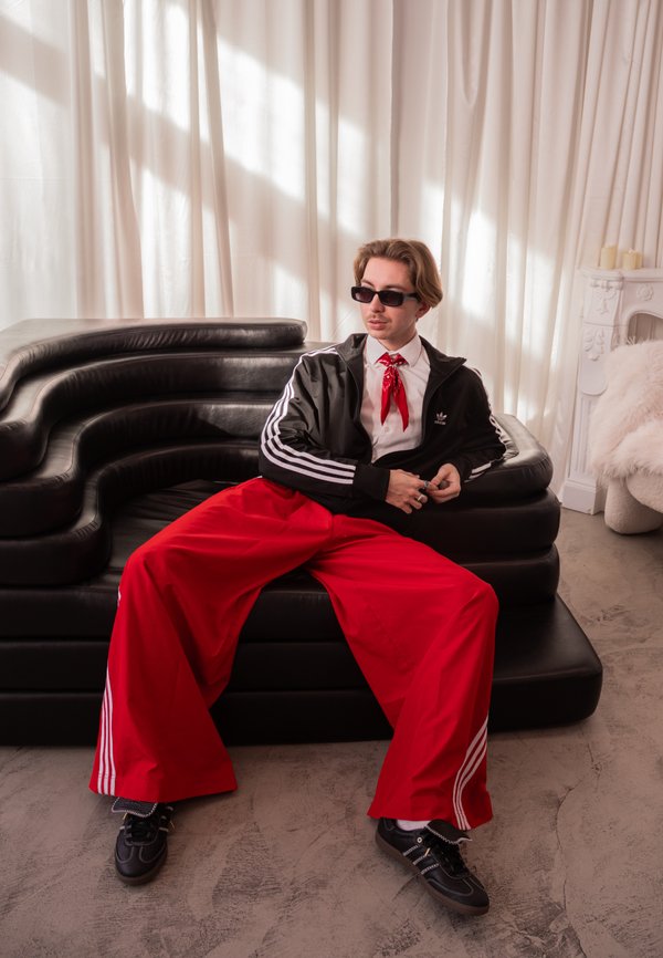 Giovane uomo con occhiali da sole, giacca nera, pantaloni rossi ampi e sciarpa rossa al collo seduto su un divano moderno nero in una stanza luminosa con tende bianche.