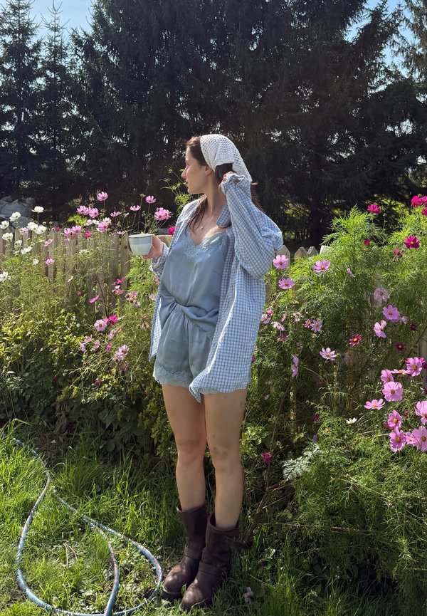 Femme en tenue bleue et foulard sur la tête tenant une tasse, debout près de fleurs roses et blanches dans un jardin ensoleillé avec des arbres persistants foncés derrière.