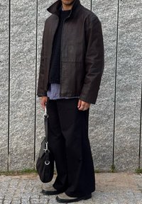 Κ αφέ δερμάτινο μπουφάν με ψηλό γιακά πάνω από μια μαύρη υφασμάτινη μπλούζα, συνδυασμένο με φαρδιά μαύρα παντελόνια και γκρίζα παντόφλες. Μαύρη τσάντα.