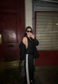 Top preto sem mangas, blazer oversized preto, calças pretas com riscas brancas, óculos de sol pretos, segurando uma bolsa escura, em frente a uma porta vermelha.