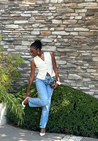 Λευκό αμάνικο γιλέκο με ένα κουμπί, ανοιχτό μπλε τζιν, λευκά μυτερά τακούνια και μια μαύρη τσάντα, ποζαρισμένο μπροστά σε έναν πέτρινο τοίχο.