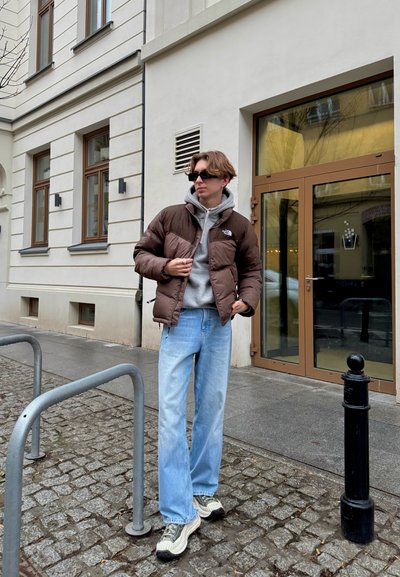 Chaquetón marrón, sudadera gris, jeans azul claro, zapatillas con patrón verde y blanco. De pie junto a barandillas metálicas grises en una calle de adoquines.