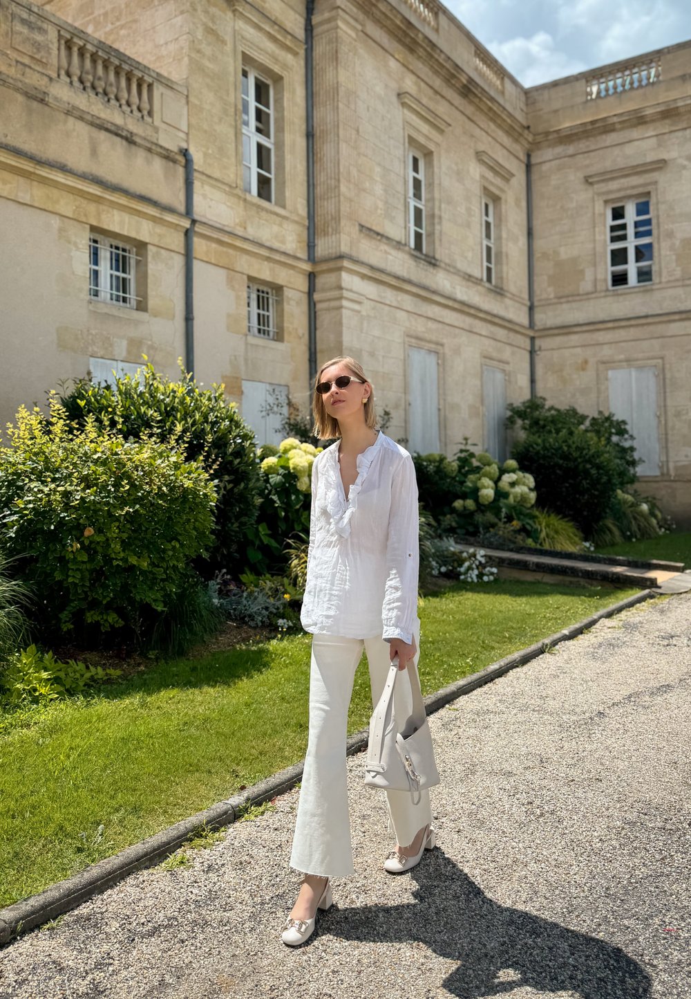 Blouse en lin blanc avec un col volanté, assortie à un pantalon flare blanc. Un sac à main gris clair et des chaussures beiges complètent l'ensemble. Verdure en arrière-plan.