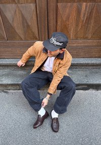 Uomo con berretto nero, giacca marrone chiaro, camicia bianca, pantaloni neri larghi e scarpe marroni seduto su gradini di pietra mentre sistema la gamba dei pantaloni.